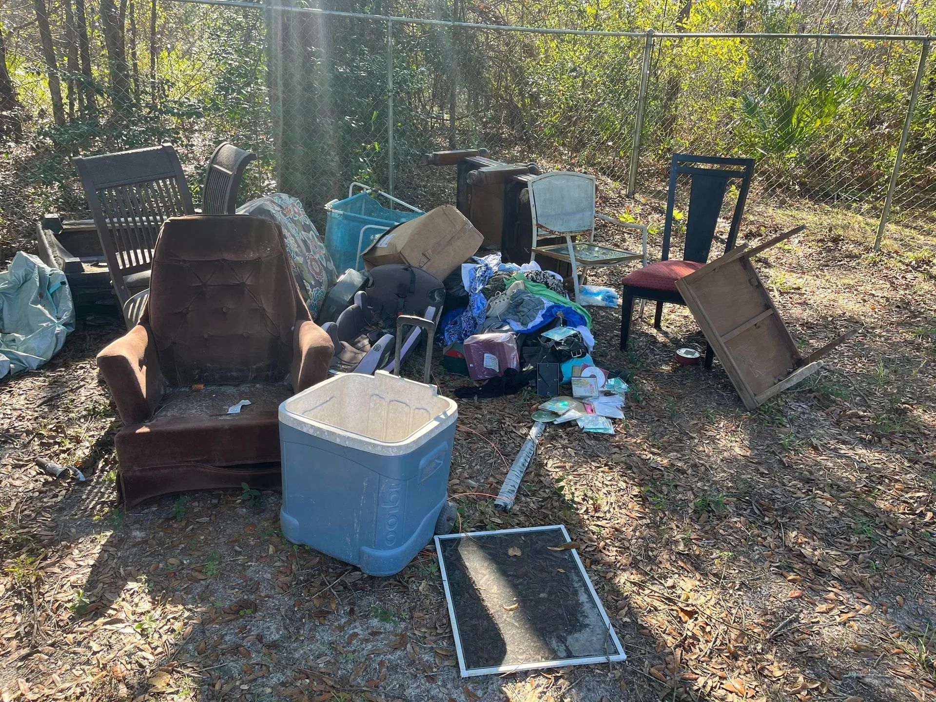 Pile of discarded furniture and trash near a chain-link fence, outdoors.