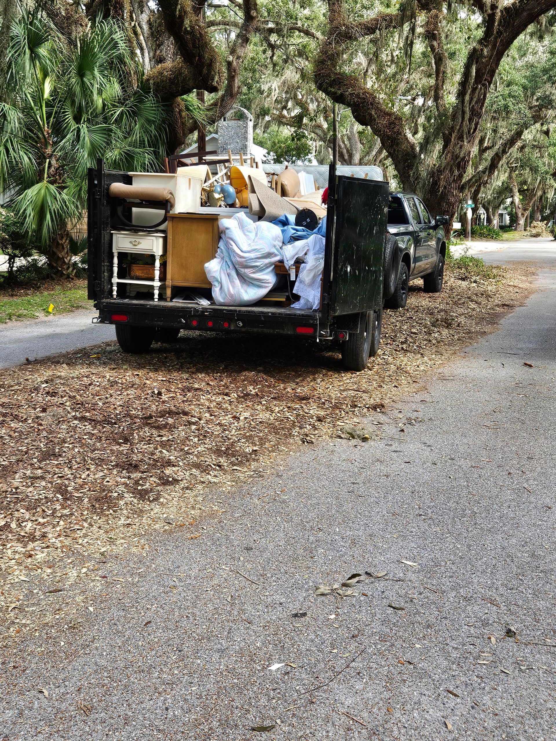 Truck bed filled with furniture and items on a gravel road covered in leaves, under large trees.