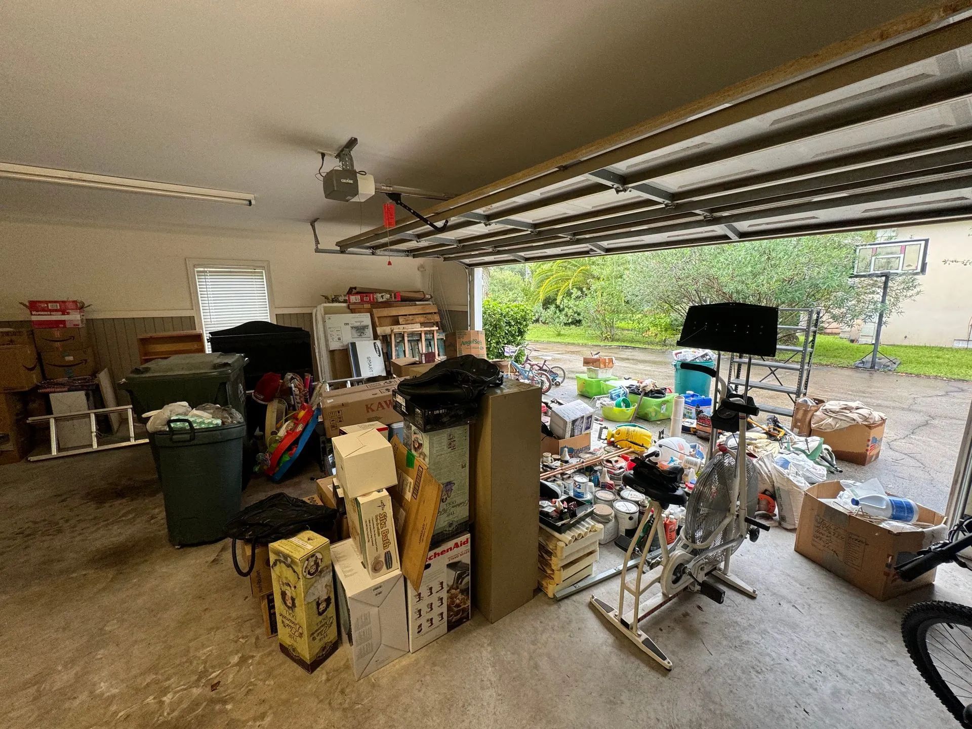 Cluttered garage with boxes, equipment, and trash cans; open garage door with basketball hoop in view.