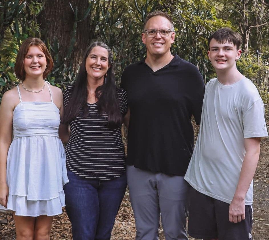 Family of four smiles for a photo outdoors. Green trees form the background.