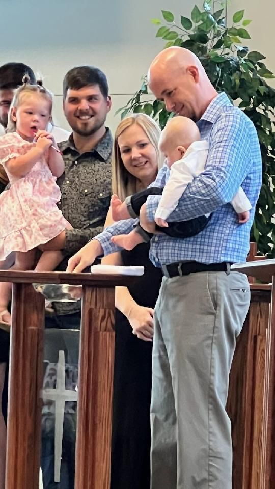 Man holding a baby prepares for a baptism. Others watch, inside a church-like building.