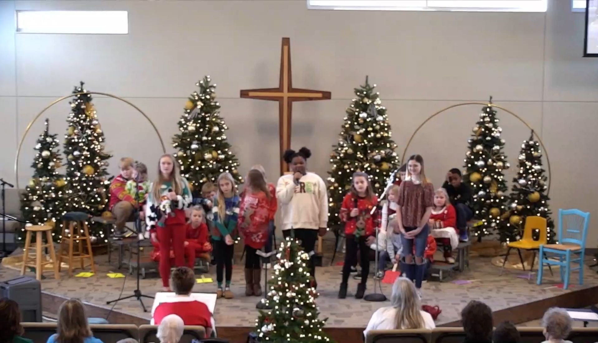 Children singing on stage with Christmas decorations and a cross.