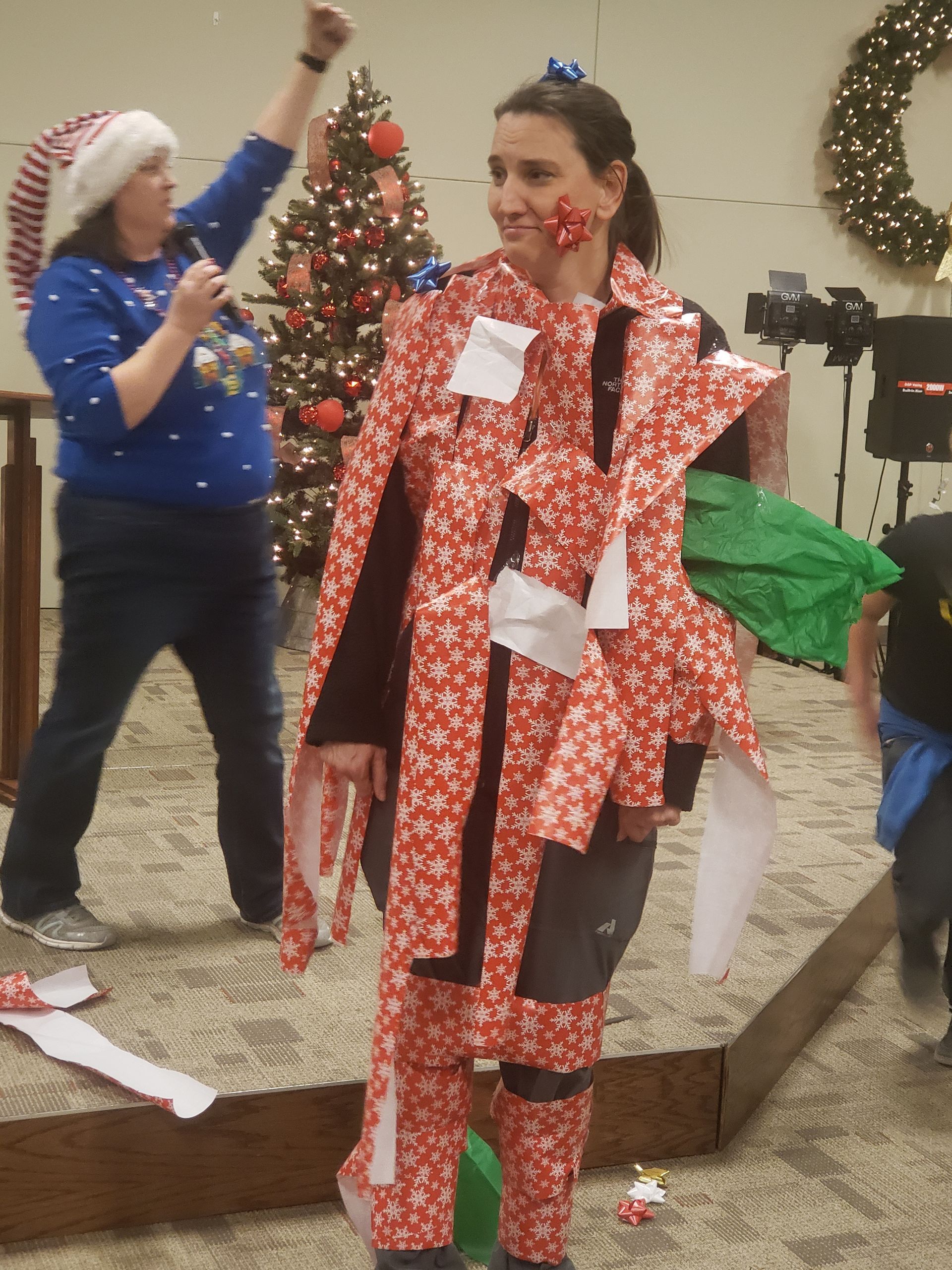 Woman covered in wrapping paper stands on stage, another woman with a microphone gestures. Christmas decorations in the background.