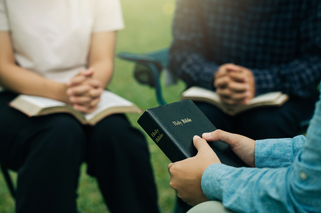 Person holding a Bible, seated with two others who are also holding books, outdoors.