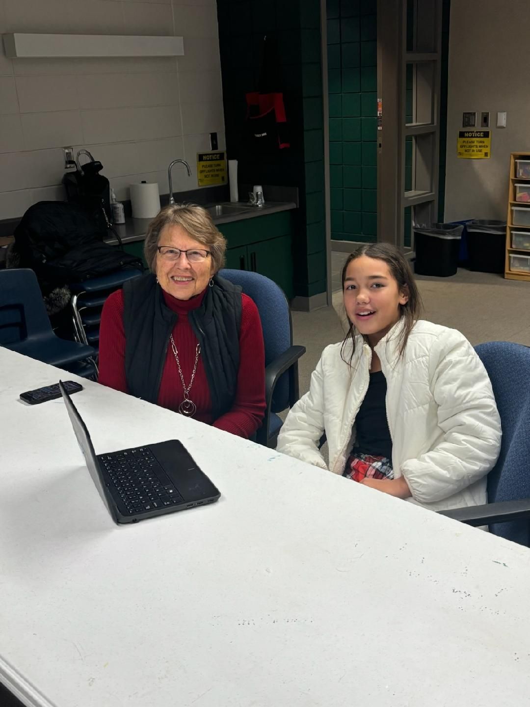 Two people at a white table. Older woman in red and black, smiling. Young person in white jacket, smiling. Laptop.