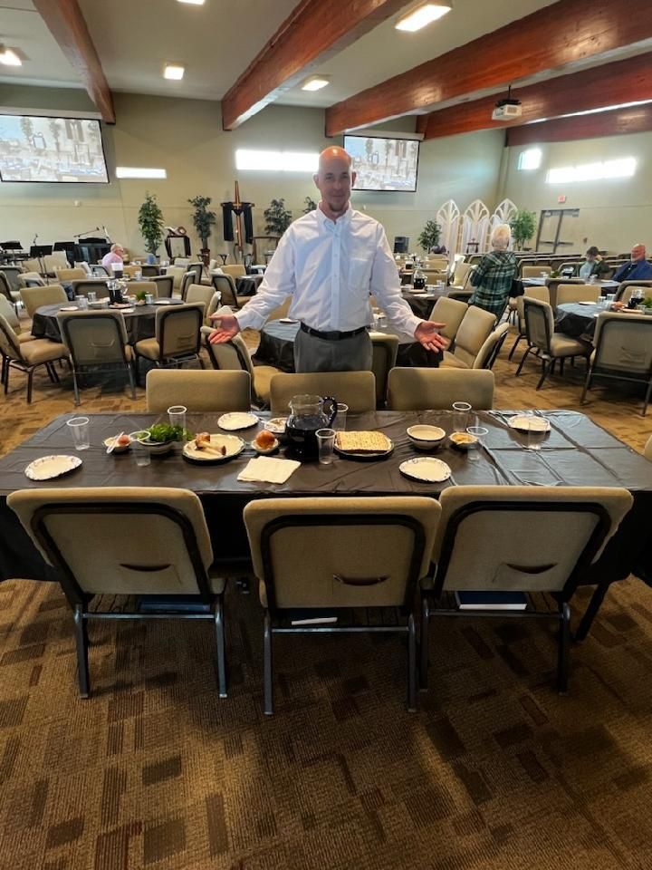 Man gestures in a dining hall set for a meal. Empty tables and chairs. Overhead TVs.