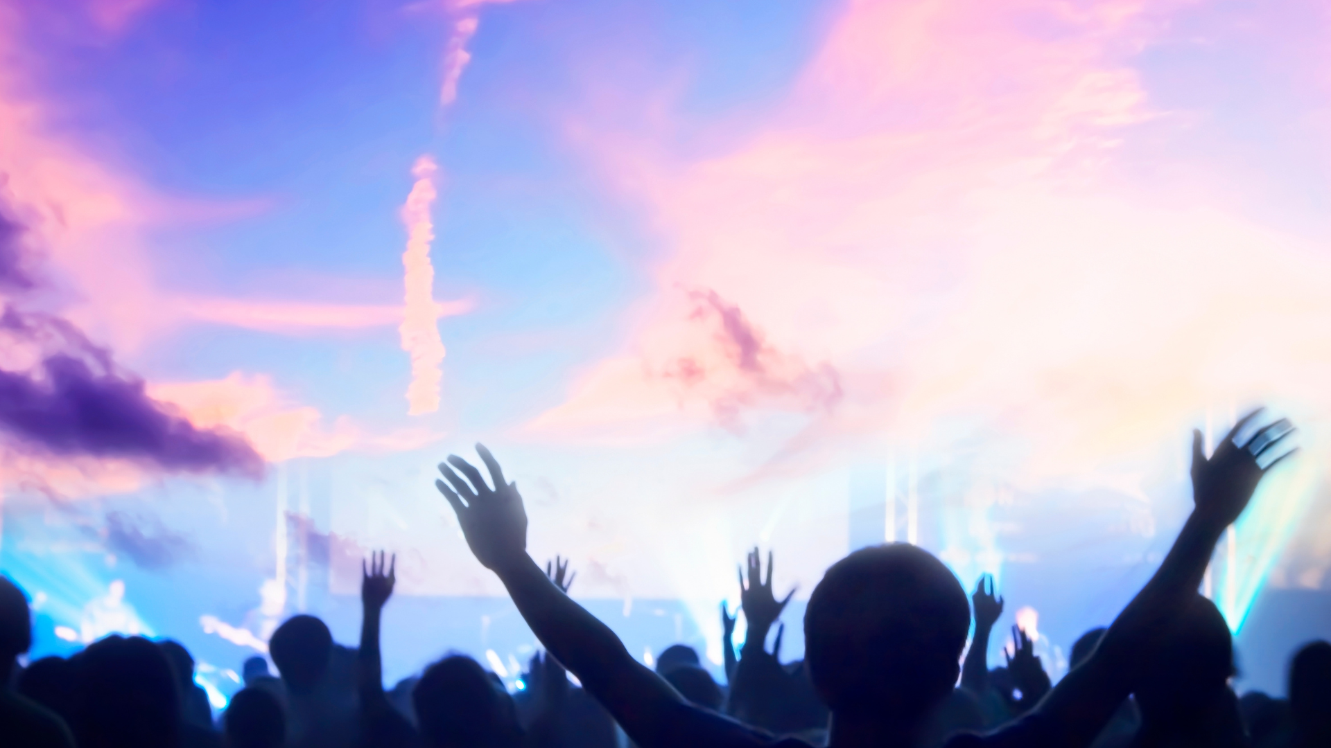 People with raised arms facing stage with bright sky, worship or concert setting.