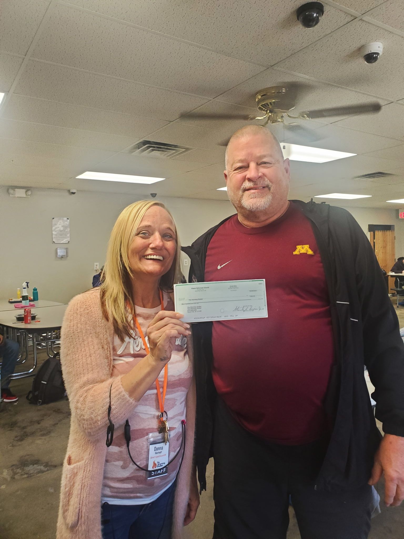 Woman and man holding a check; woman smiles. Indoors, people and tables in background.
