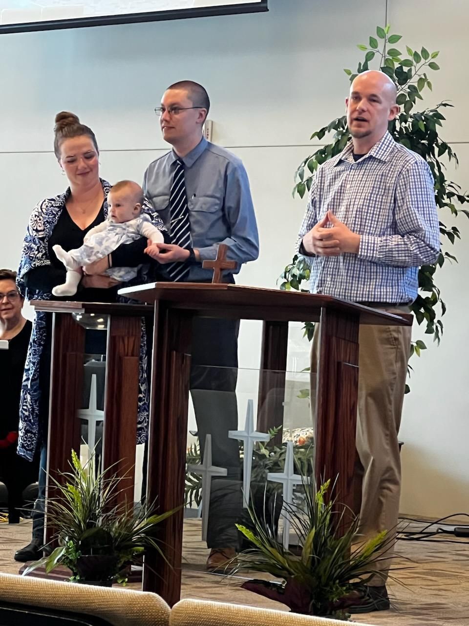 A baby is held by a woman and man at a podium with another man speaking in a church.