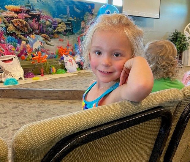 Blonde girl smiles, resting arm on chair, in front of a colorful underwater-themed backdrop and decorations.