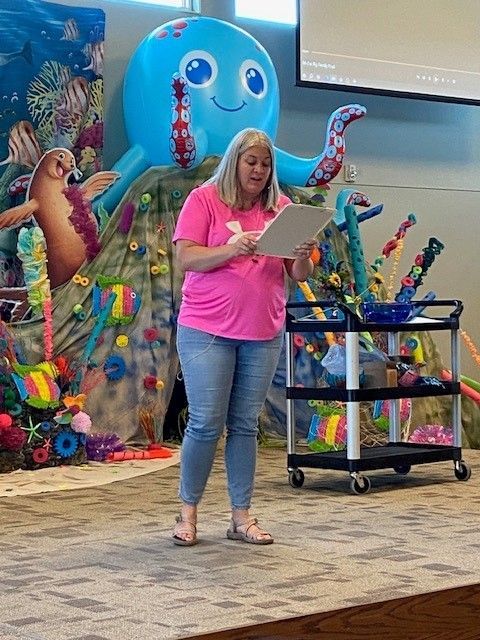 Woman in pink shirt reads from paper in front of underwater-themed decorations.