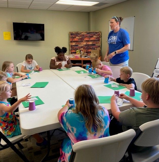 Children and an adult seated around a table, engaged in a craft project in a classroom setting.