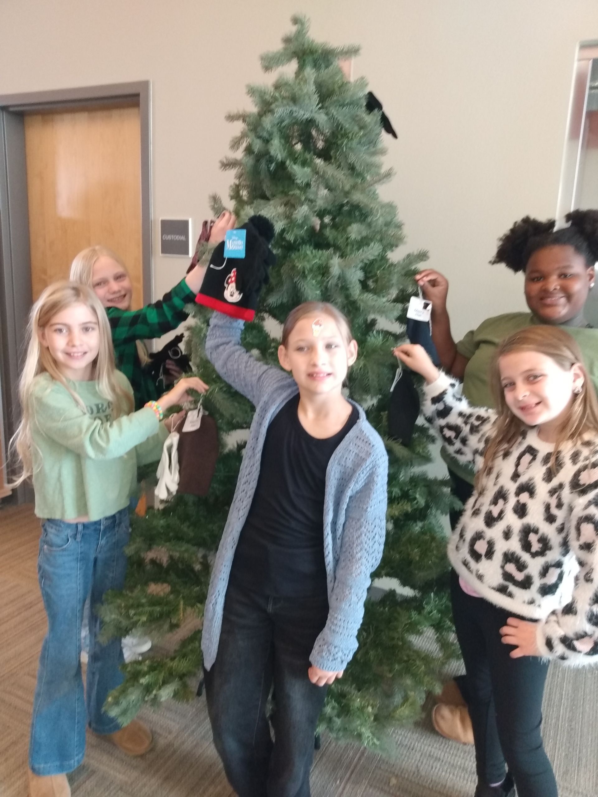 Five children holding beanies in front of a Christmas tree in a room.