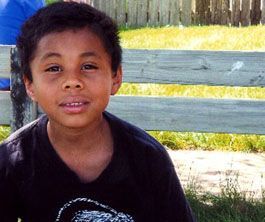 Young person smiling, outdoors in front of a wooden fence and grass, wearing a dark t-shirt.