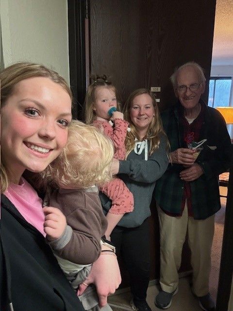 Group of five people, smiling, posing near a doorway. Two hold children. Door is brown.