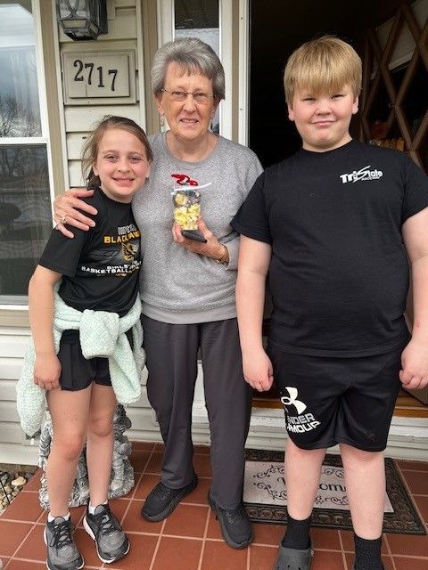 Woman holds cookies with two smiling children at a house entrance.