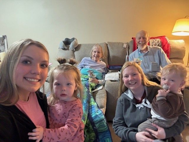 Family gathers for a photo indoors, including two women holding children, and two older adults on a sofa.