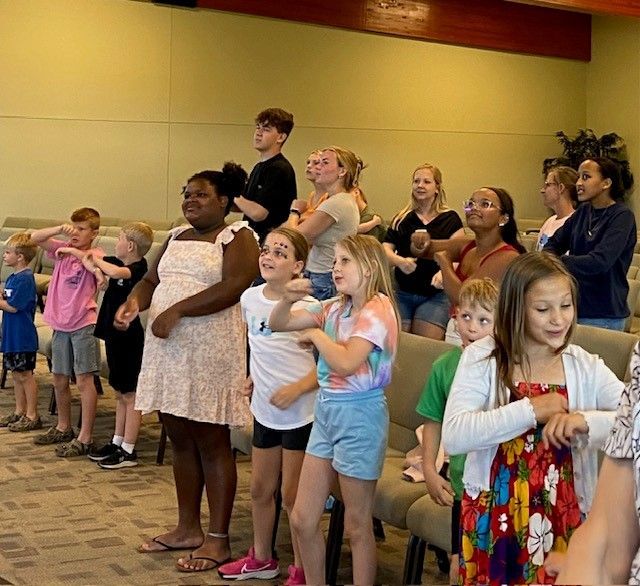 Children in a group, some standing, some seated, with arms raised indoors, possibly singing or participating in a lesson.