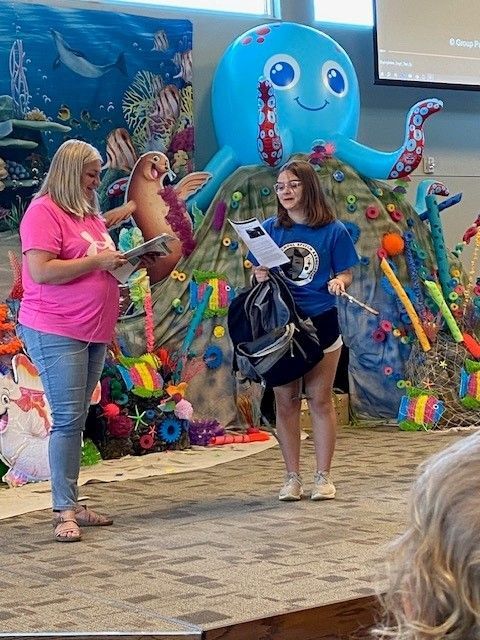 Two women stand on a stage with an ocean-themed backdrop, reading papers. One wears pink, the other blue, holding a bag.