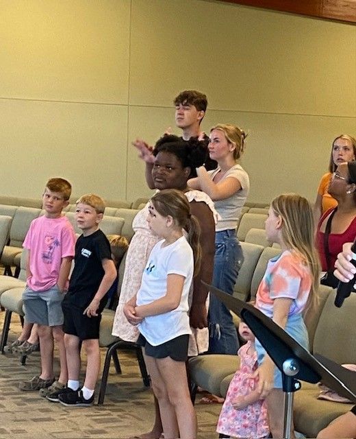 Children and adults in a church, possibly singing or listening. Some have attentive expressions.