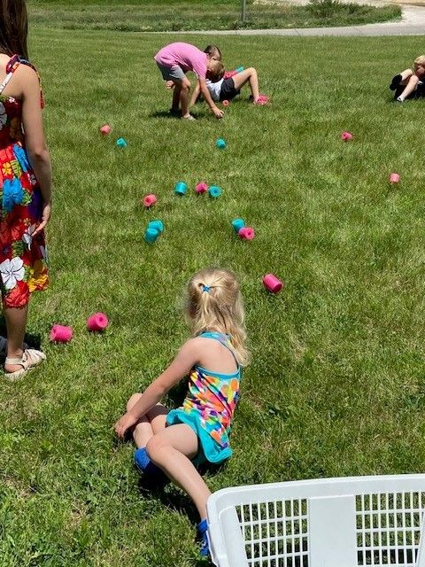 Children playing a game on green grass with pink and blue objects. One child sits, others reach and bend.