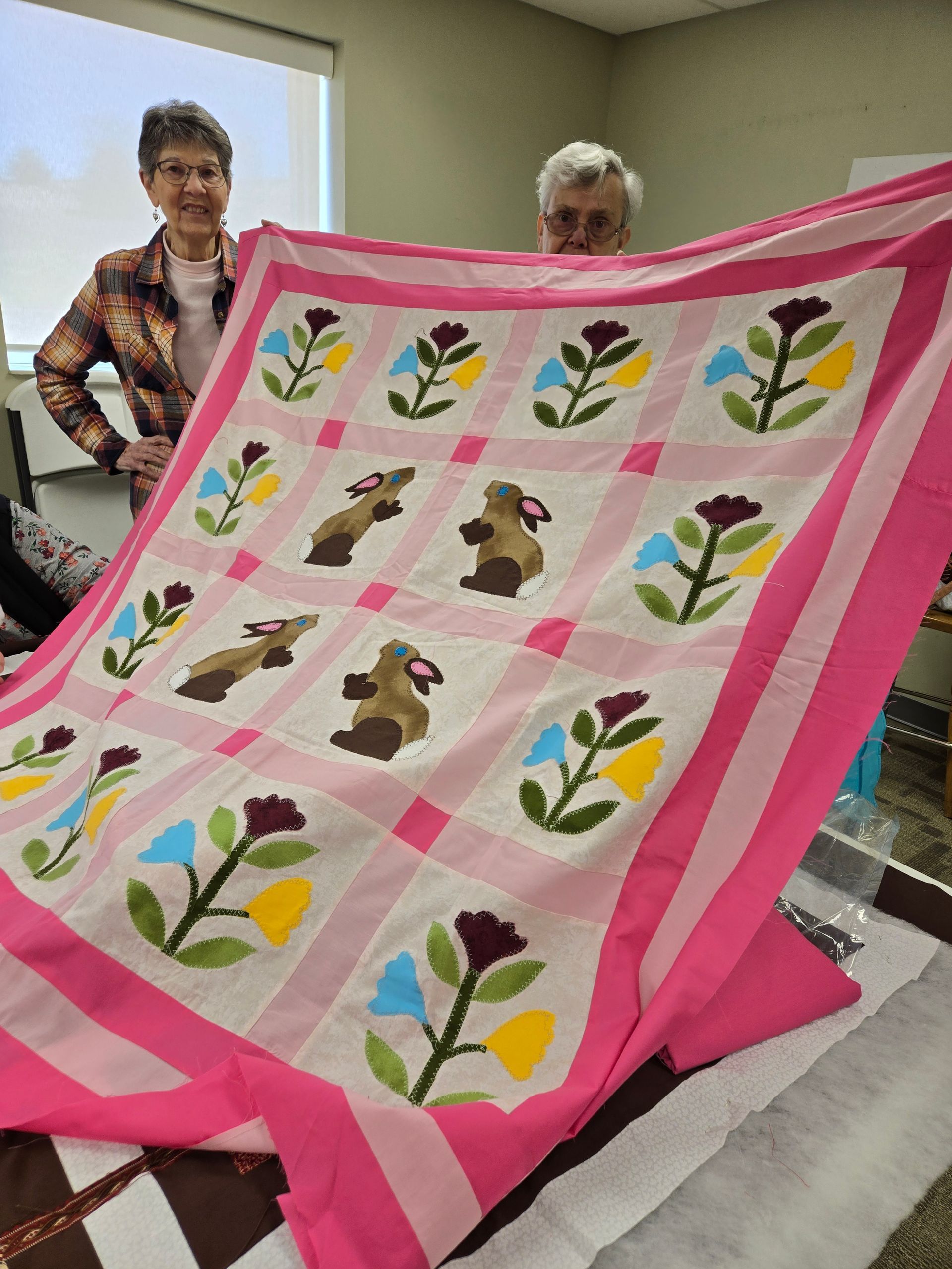 Two people holding a pink quilt with floral and animal appliqués.