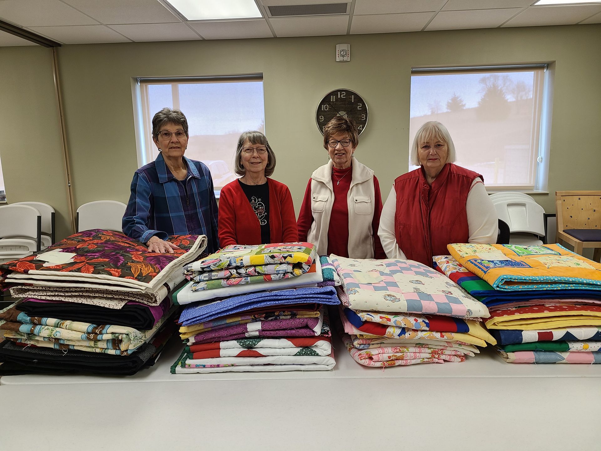 Four people stand behind stacks of colorful quilts on a table. Windows and a clock are in the background.