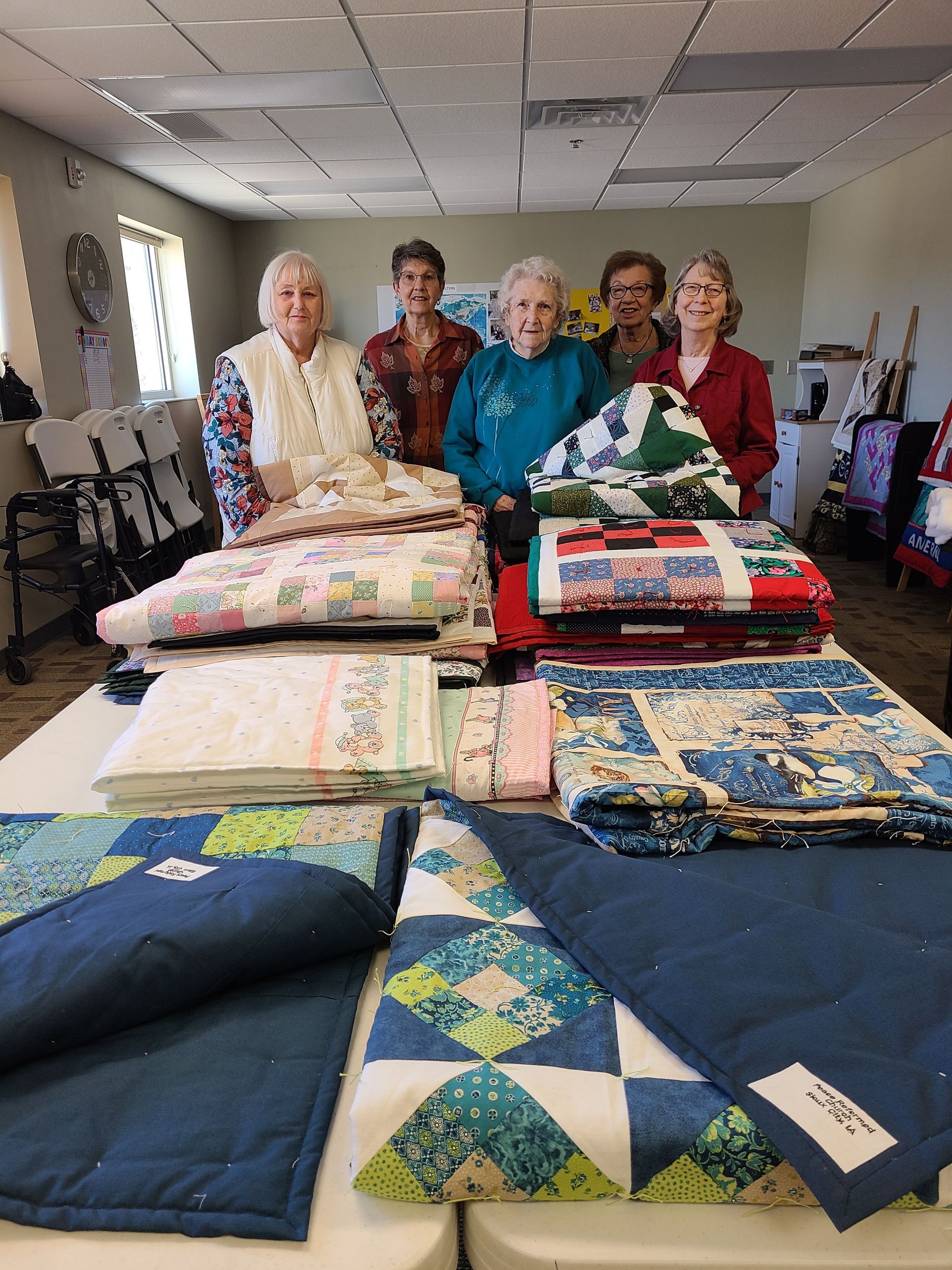 Five people stand behind a table laden with colorful quilts. They smile, posing in a room with chairs and windows.