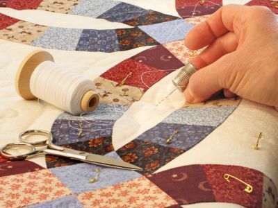 Person hand-sewing a quilt with a thimble, thread spool, scissors, and safety pins on a patchwork design.