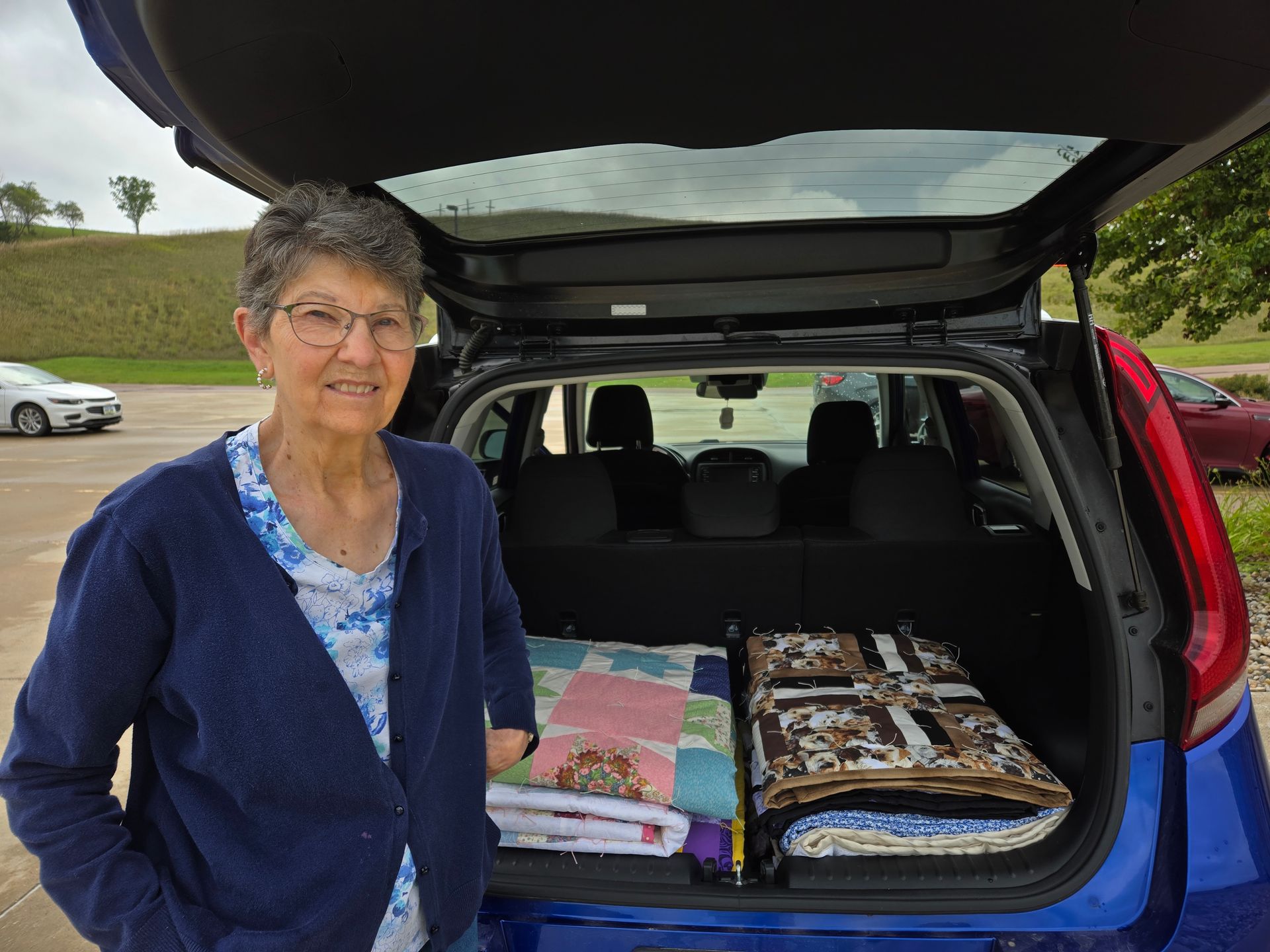 Woman standing by open car trunk with quilts. Blue car, outdoor setting.