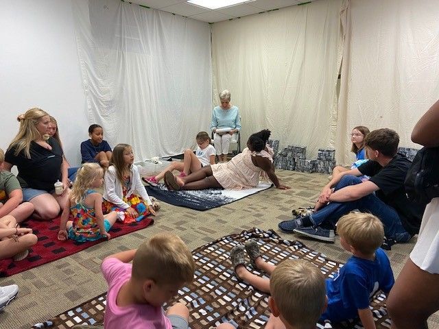 Children and an adult listen to a seated woman reading a book in a carpeted room with draped fabric walls.