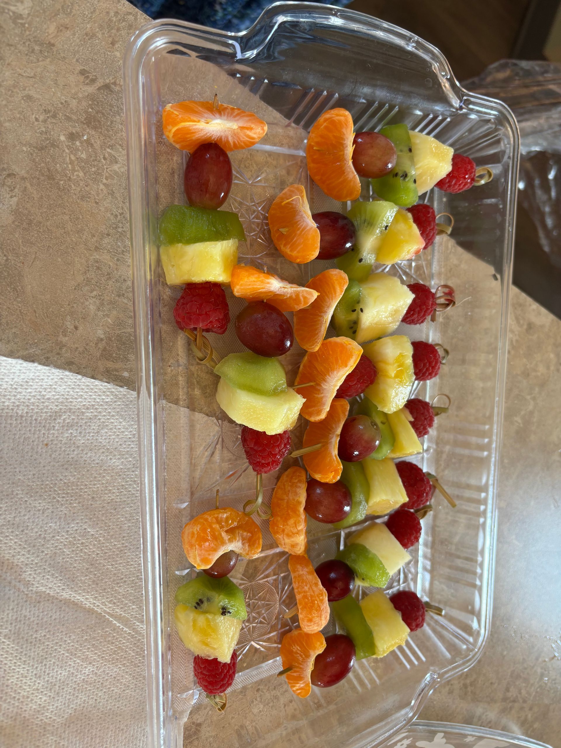 Fruit skewers in a clear rectangular dish: grapes, kiwi, pineapple, mandarin orange slices, and raspberries.