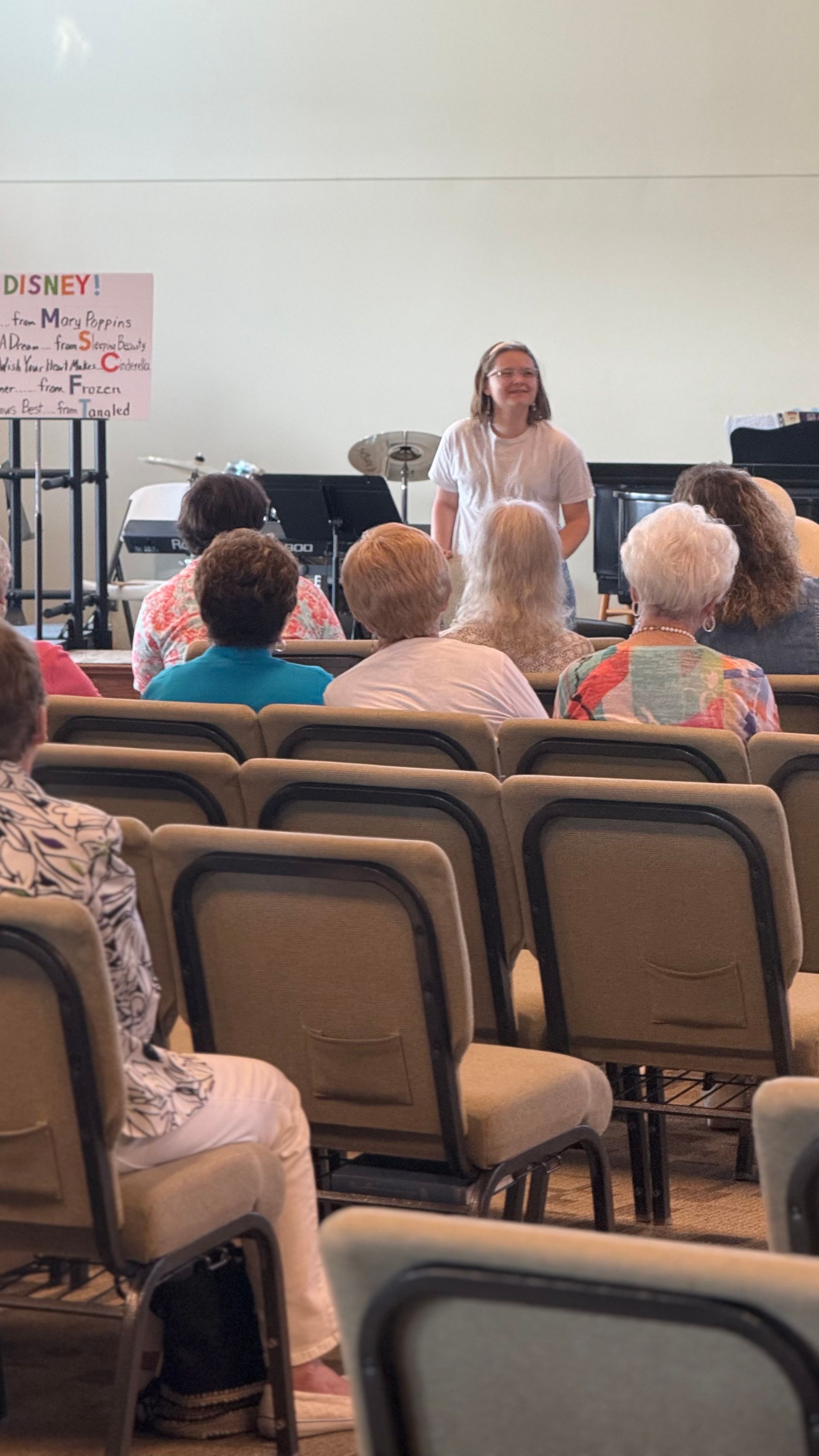 A person speaks to a seated audience. They are in a room with chairs and a music stand.