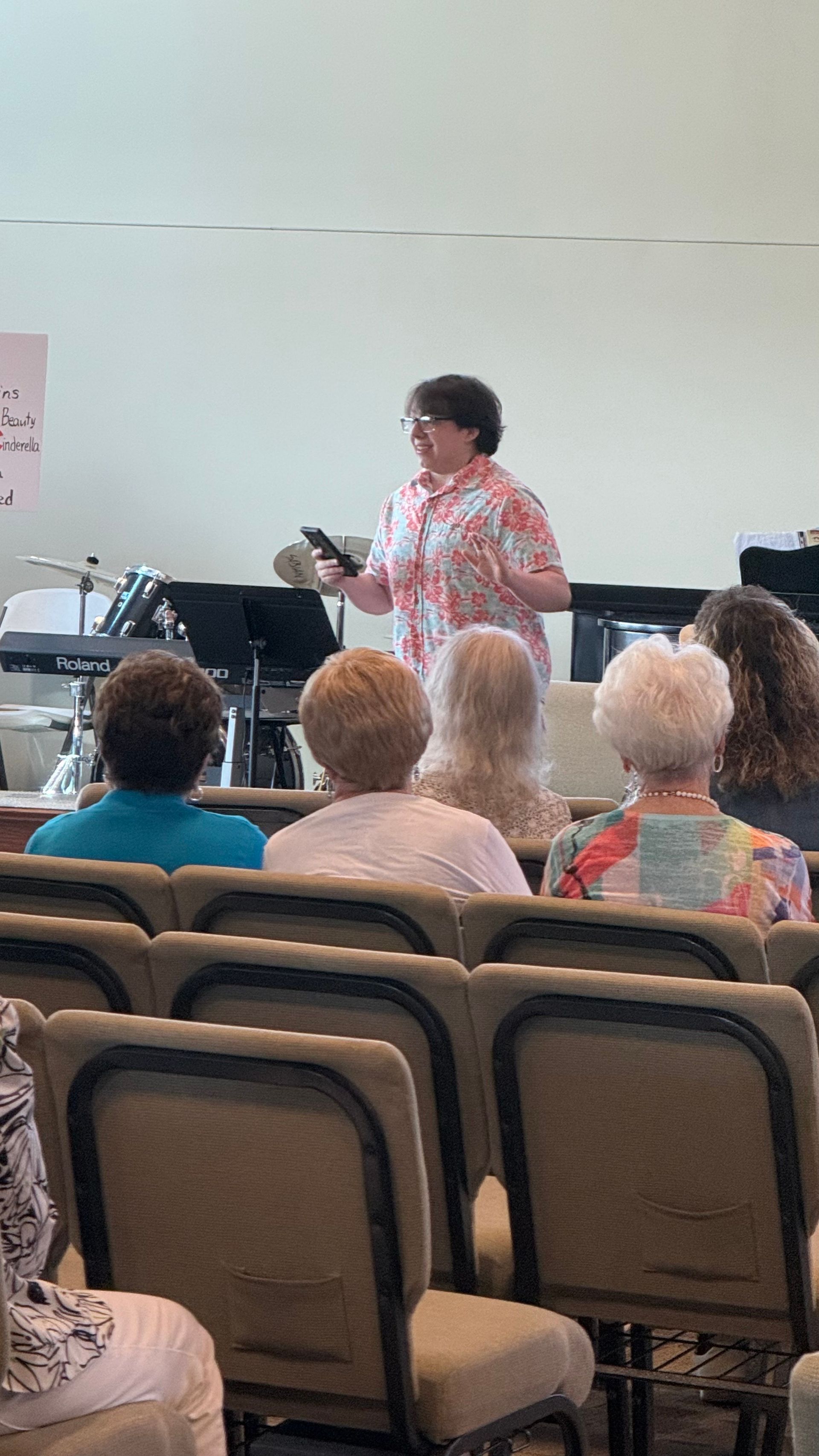 Woman speaking to an audience in a room; a musical keyboard and equipment are visible in the background.