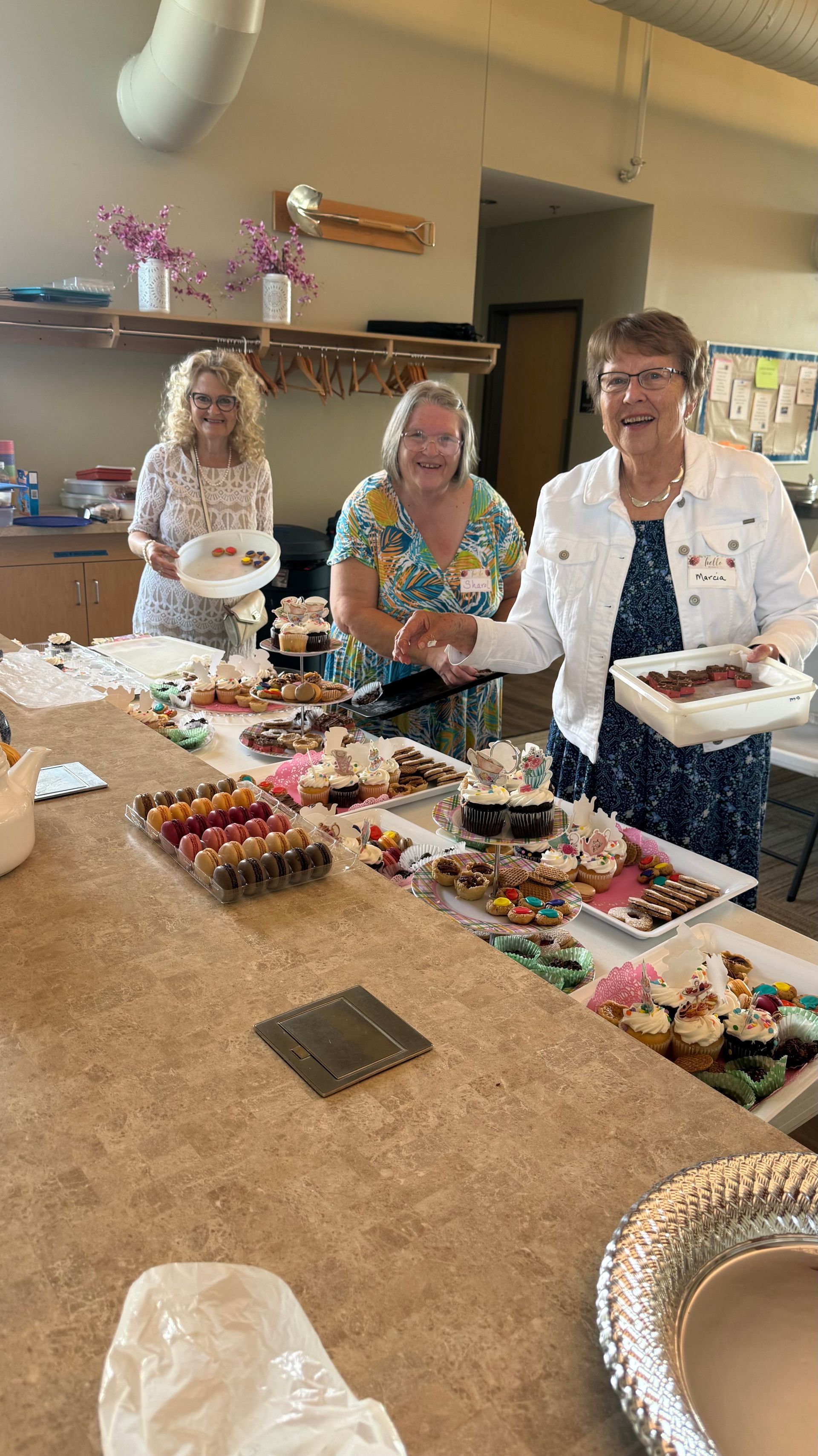 Three women at a buffet table laden with food, smiling. One holds a plate, another a tray of bacon.