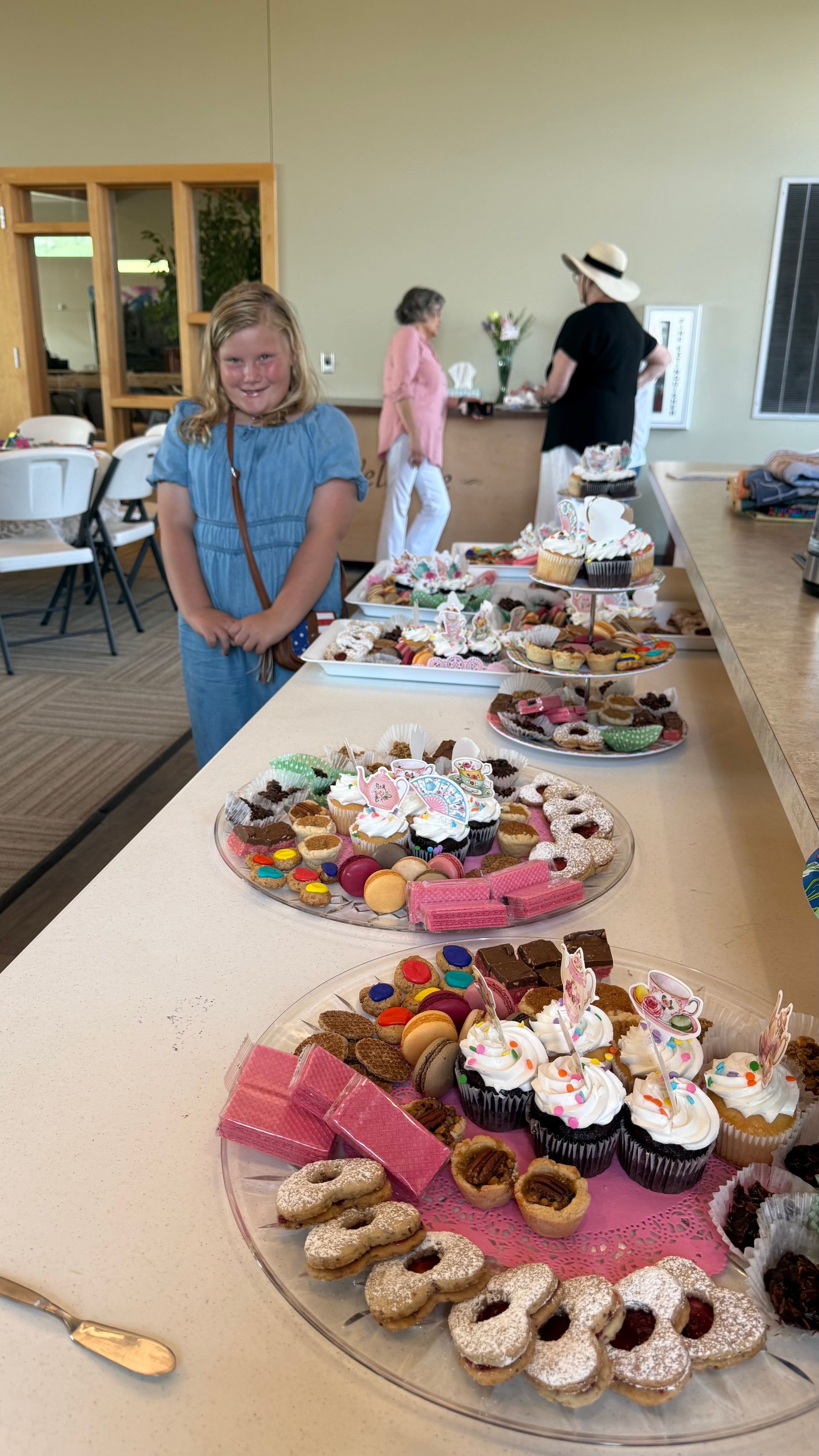 A young person smiles in front of a table laden with pastries. Others stand in a bright room.