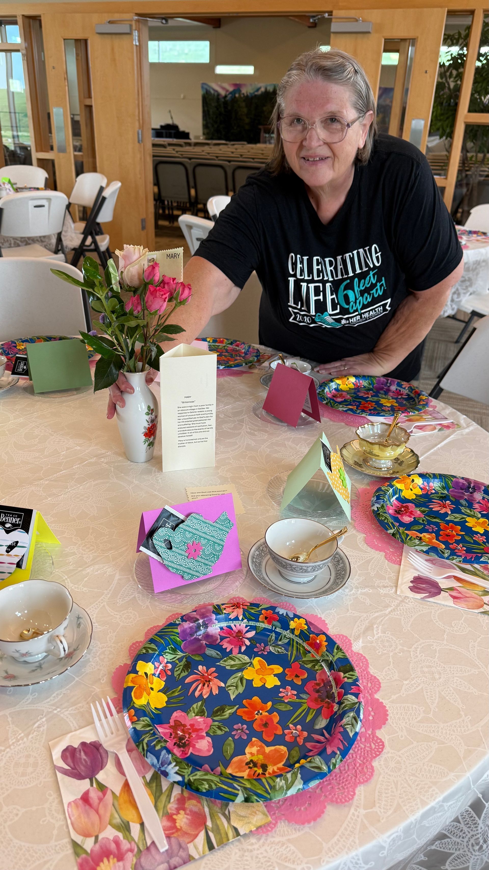Woman sets a table with floral plates, teacups, and a vase of flowers, indoors.