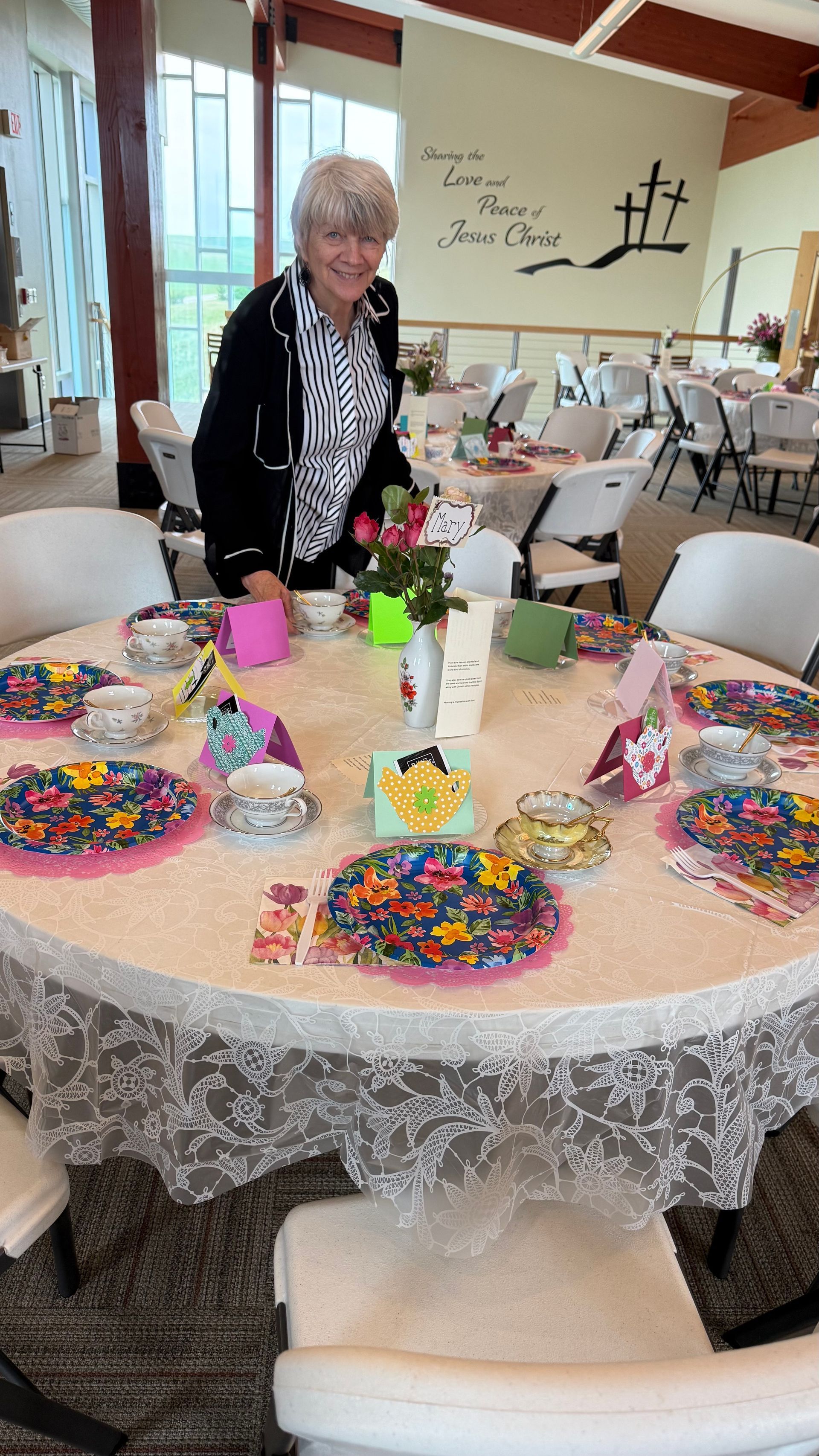 Woman setting a round table decorated for an event, floral plates and decorations, in a bright room.