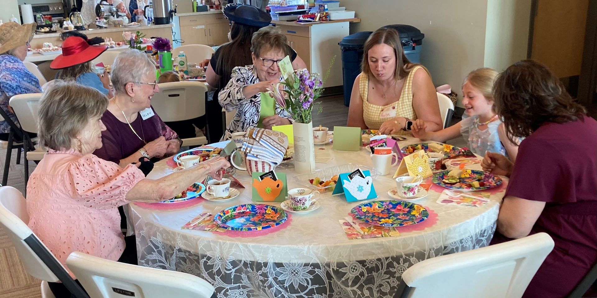 People seated around a table, enjoying a tea party. Dishes and colorful decorations are visible.