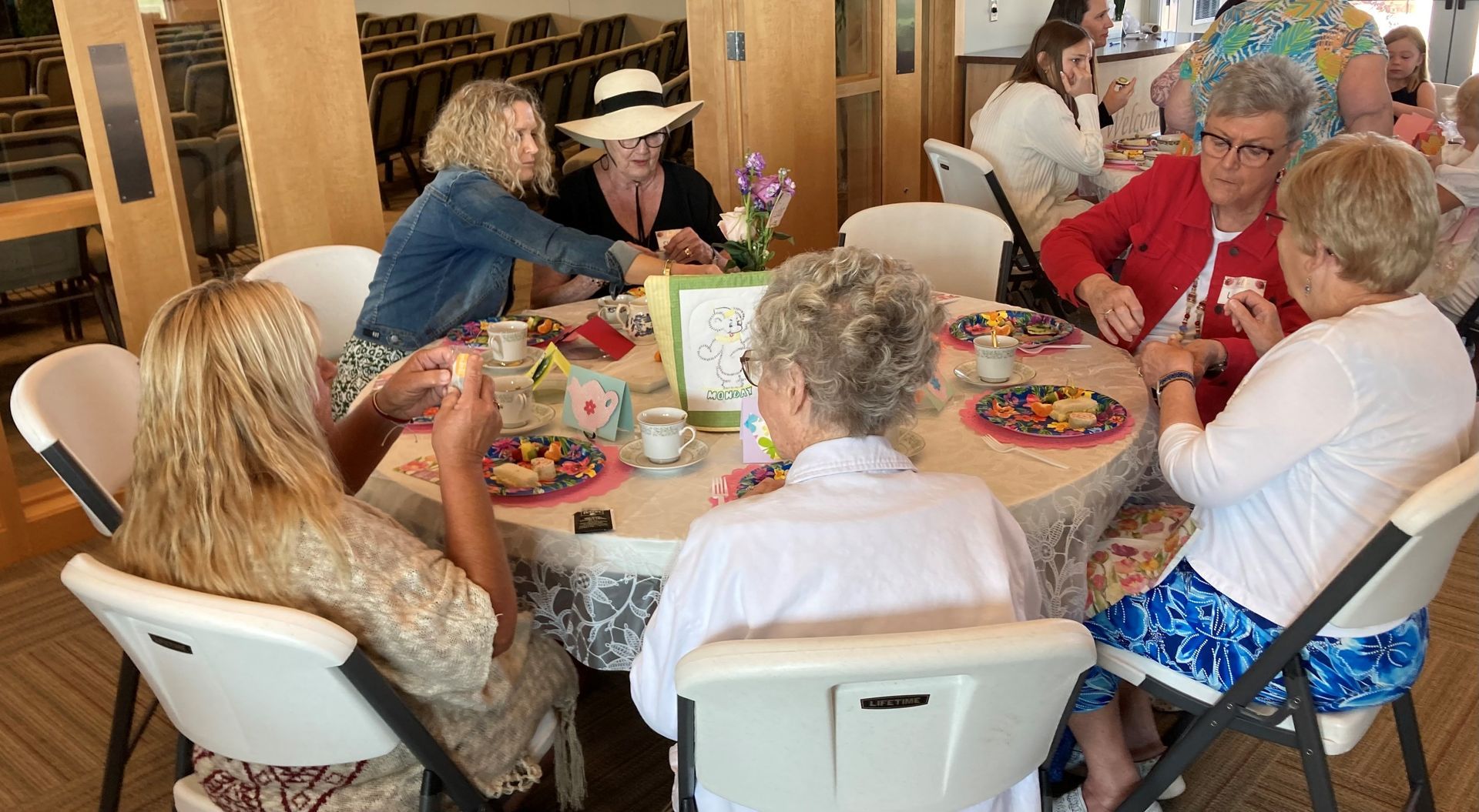 People at a round table, eating and talking. Flowers in center, room with wooden columns, sunny.