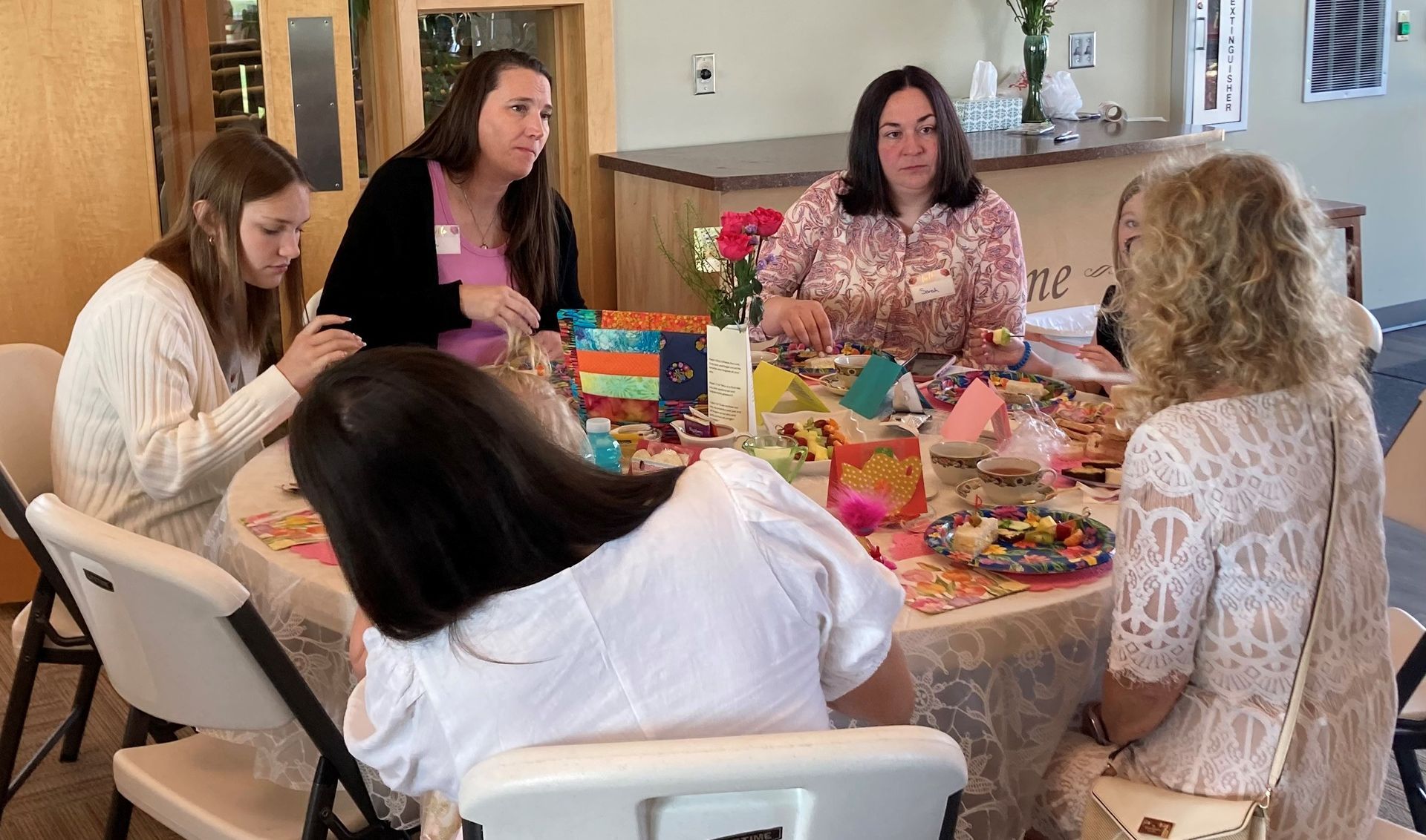 Five people seated at a round table with gifts, eating food. Bright, indoor setting.