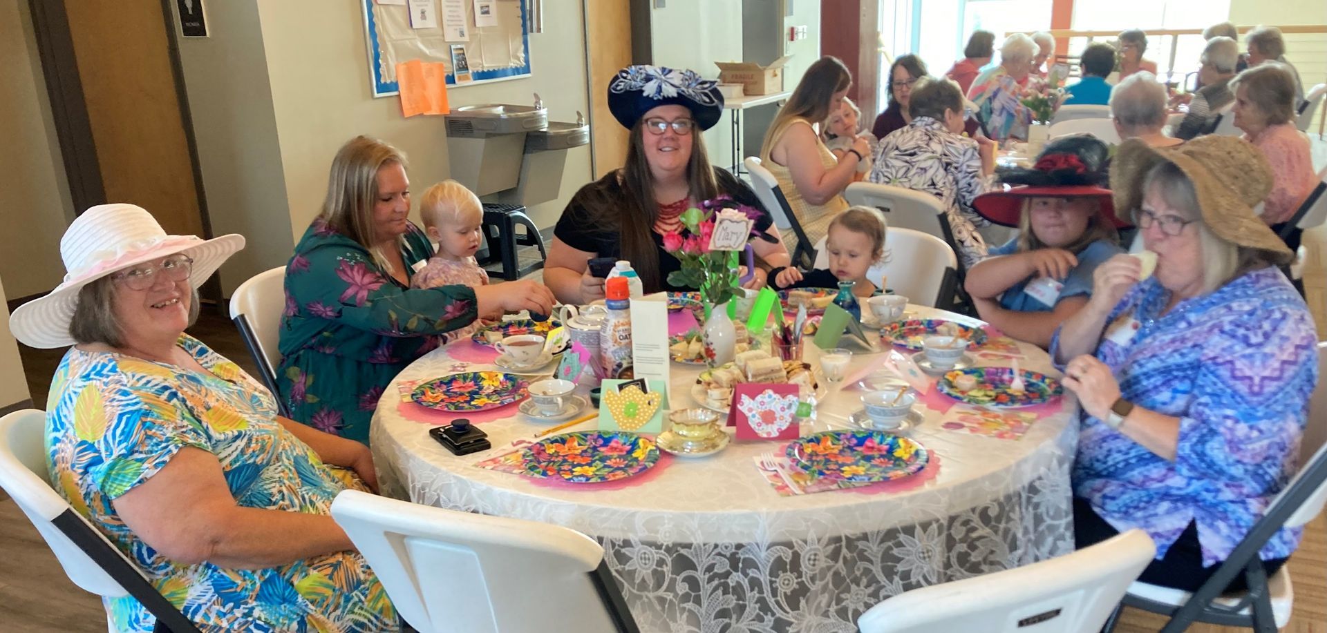 People at a tea party, wearing hats, seated around tables in a room, some with babies, having tea.