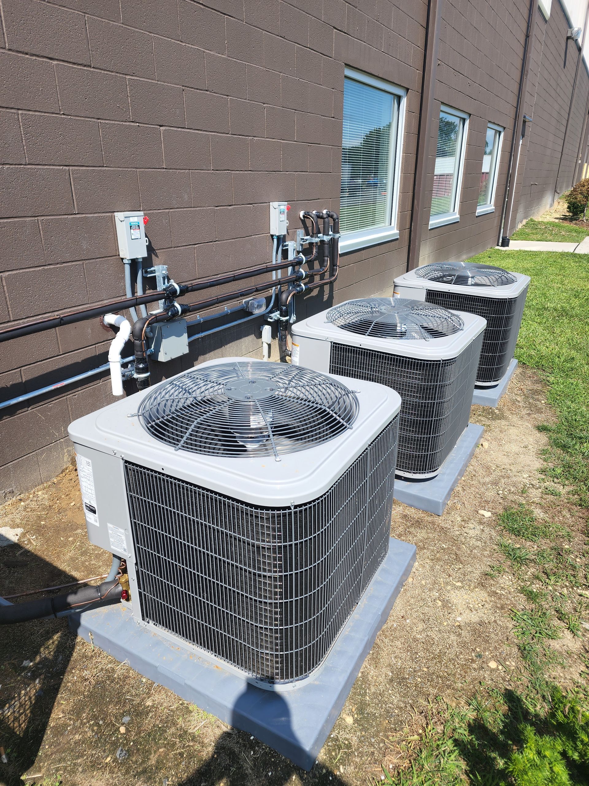 Three outdoor HVAC condenser units beside a brick building, with pipes and utility meters mounted on the wall.