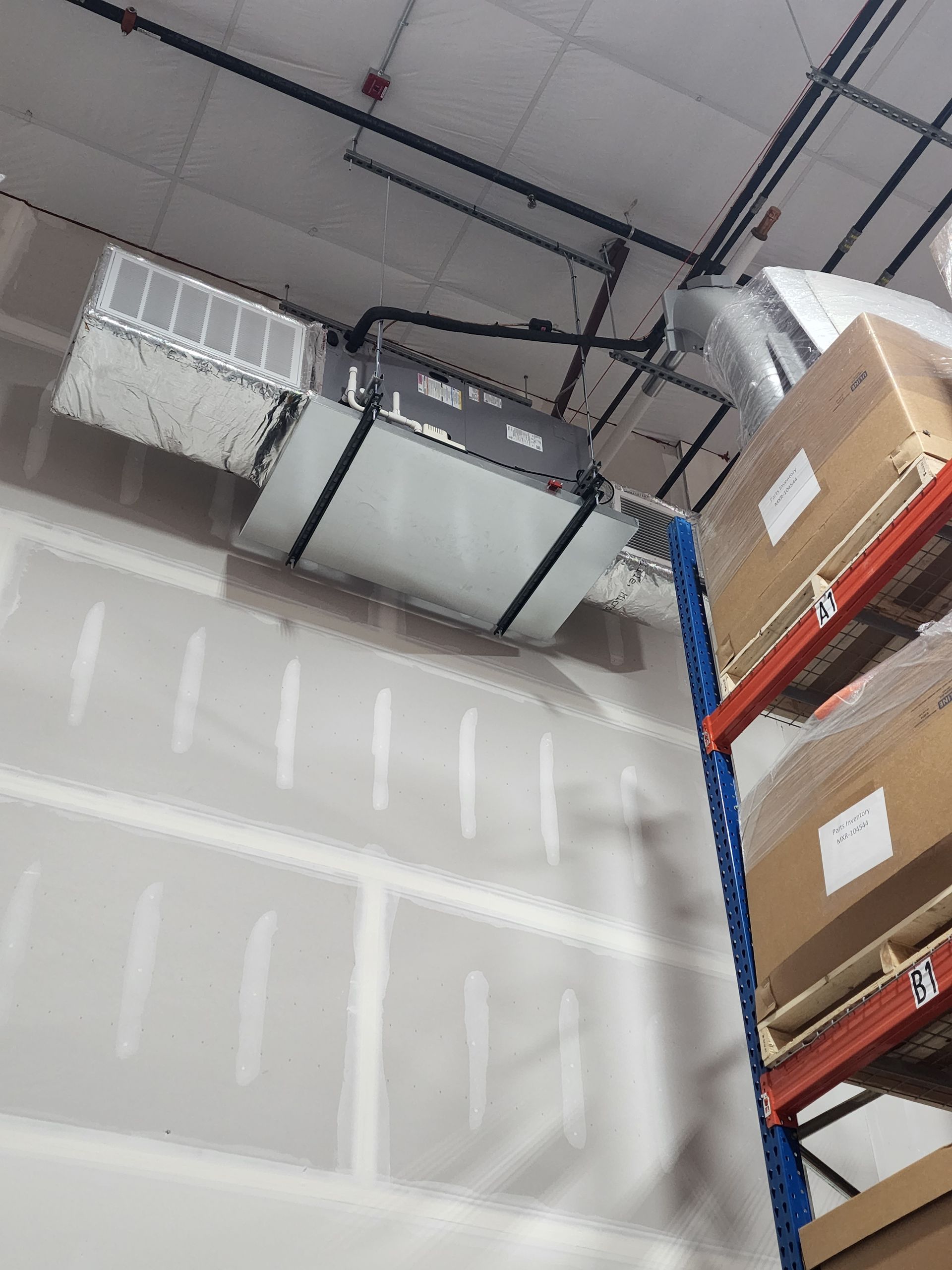 Warehouse ceiling with HVAC ducts above tall shelving stacked with cardboard boxes.