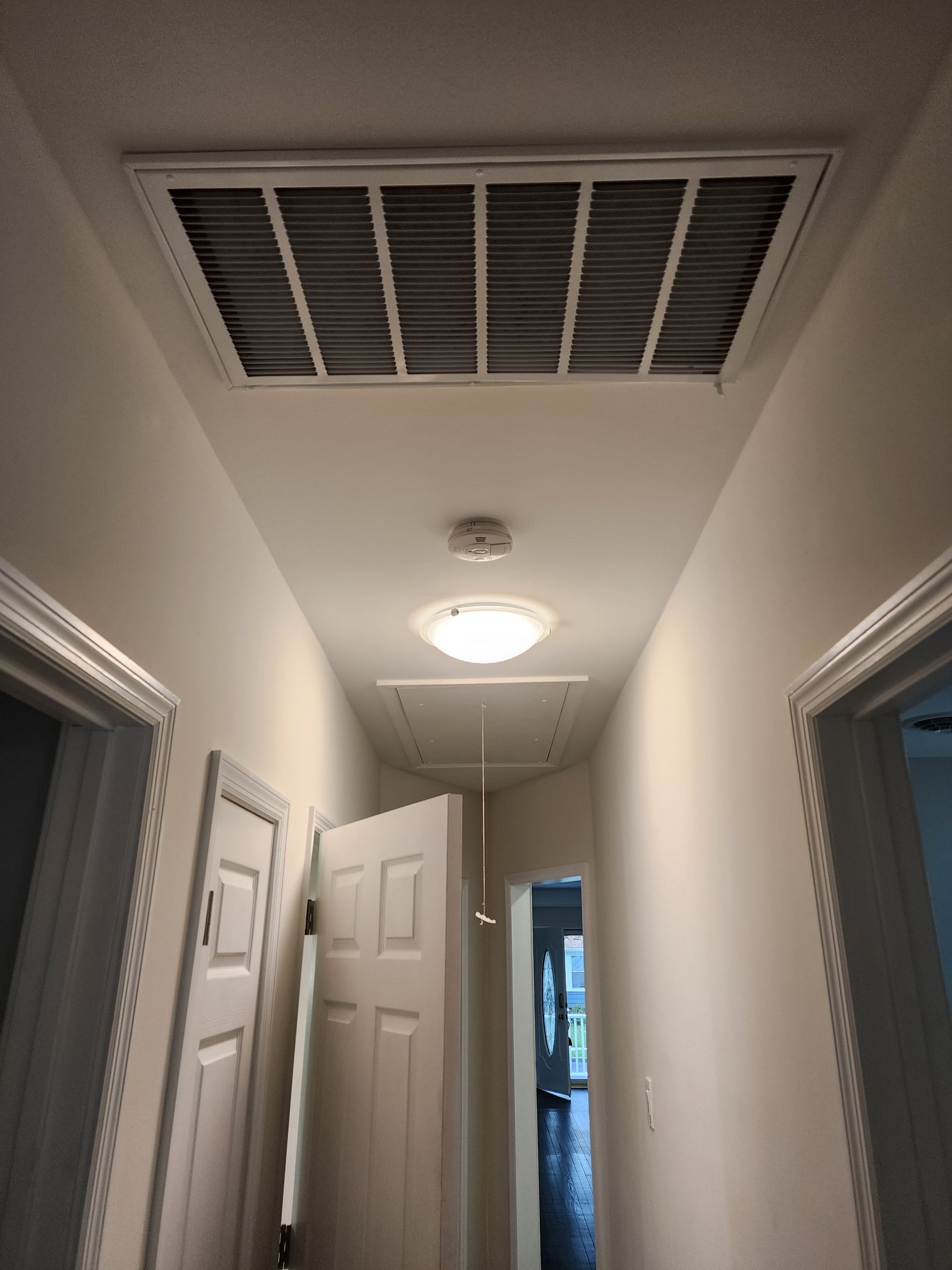 Narrow hallway with open white doors, ceiling vent and light, leading to a brighter room.