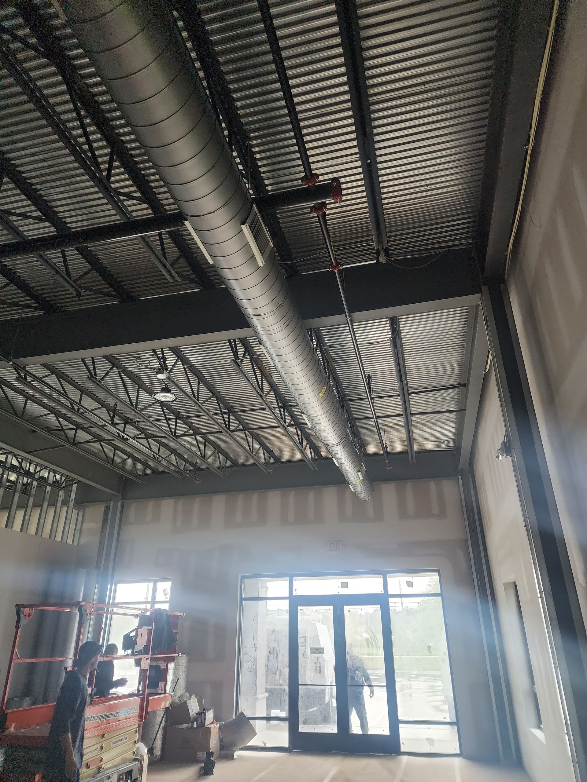 Construction site interior with exposed ceiling ducts and a red scissor lift near a glass entryway