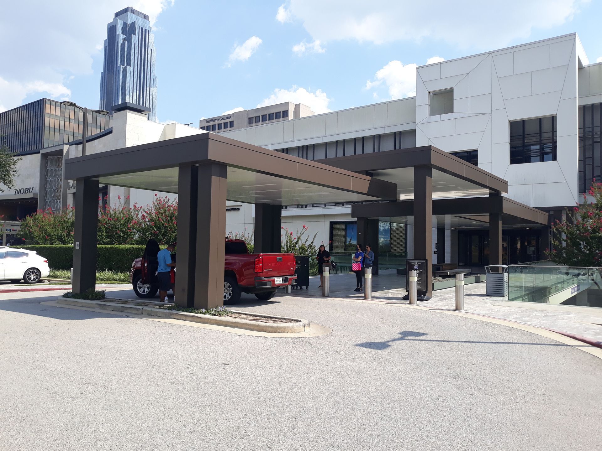 A red truck is parked under a canopy in front of a large building.
