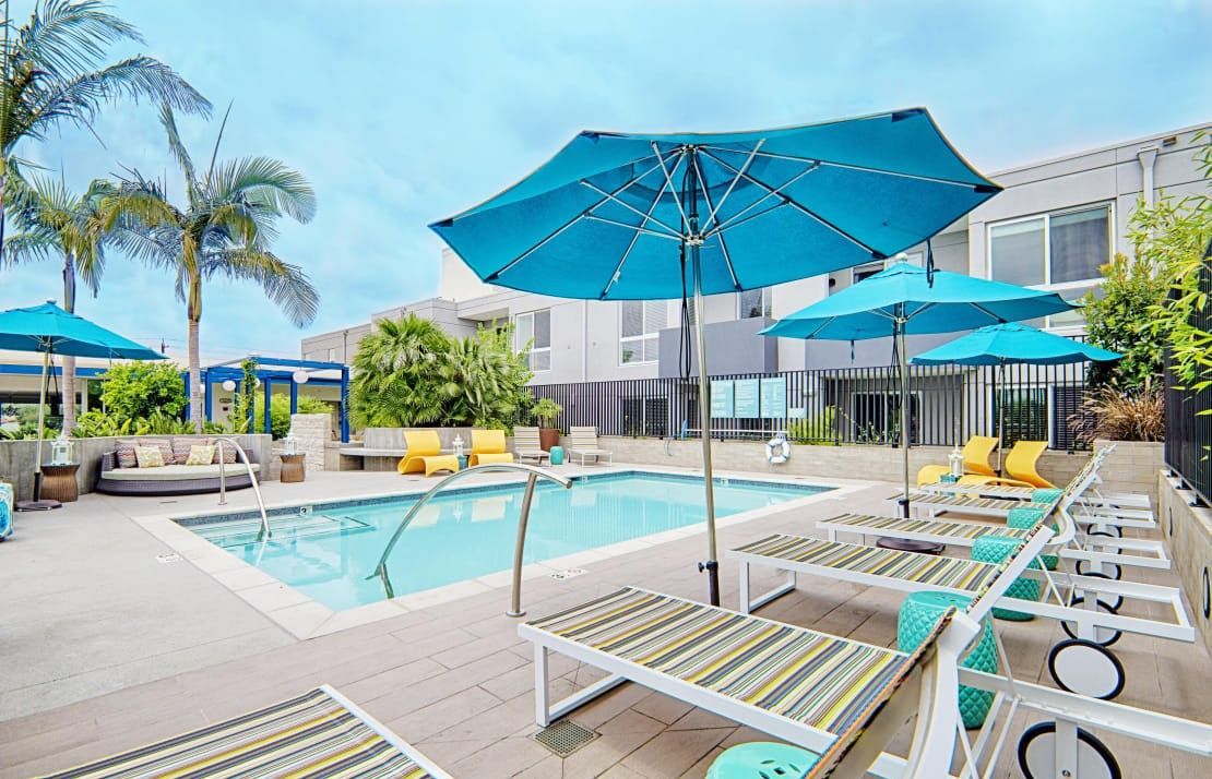 Poolside scene: turquoise umbrellas, lounge chairs, blue water, modern building, palm trees.