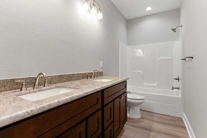 Bathroom with double vanity, tub/shower, and toilet; light gray walls, brown cabinets, and light wood-look floor.