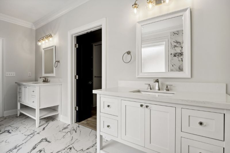 Two white bathroom vanities with mirrors and lighting fixtures, black door, marble floor.